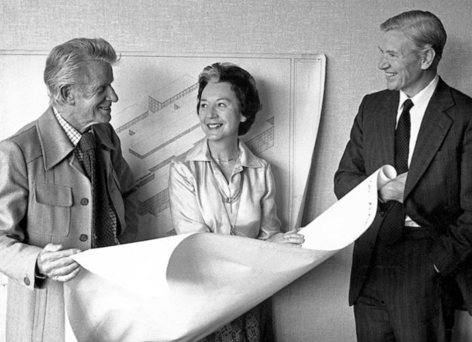 Lucienne Day and Robin Day discussing design plans with a colleague, holding an architectural blueprint.