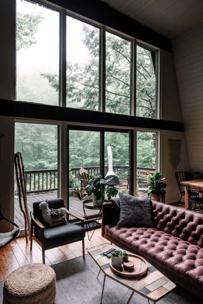  Interior of the Hunter Greenhouse featuring a cozy living room with a burgundy tufted sofa, large floor-to-ceiling windows, and lush forest views.
