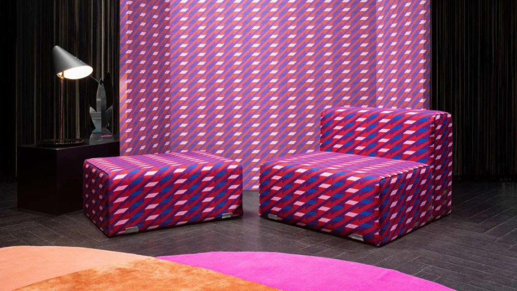 A modular furniture set designed by Kazuhide Takahama, featuring geometric-patterned upholstery in shades of pink, red, and blue. The set includes a lounge chair and ottoman, complemented by a matching fabric wall backdrop.