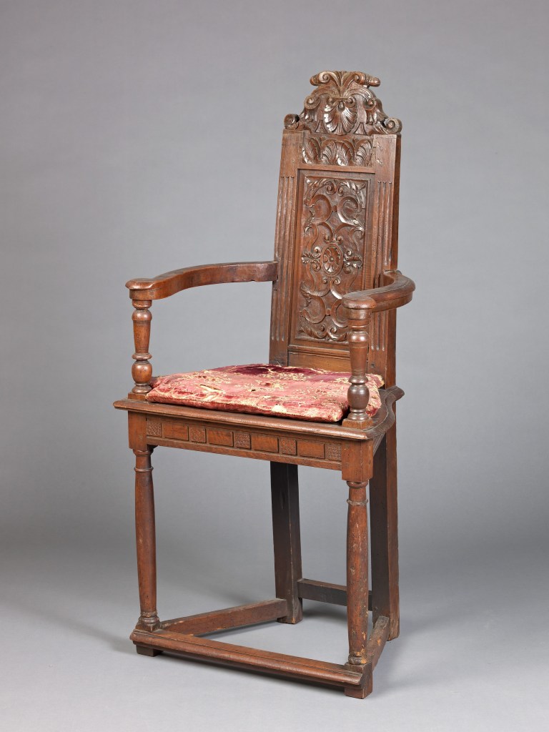 Antique French caquetoire chair with intricate wood carvings, turned arm supports, and an upholstered seat cushion.