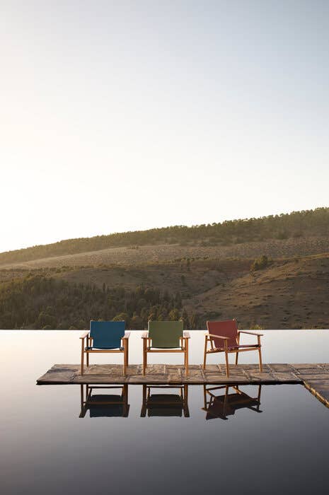 Three Cordoba Armchairs by Foster + Partners set on a floating wooden platform, overlooking a calm infinity pool with a scenic mountain backdrop.