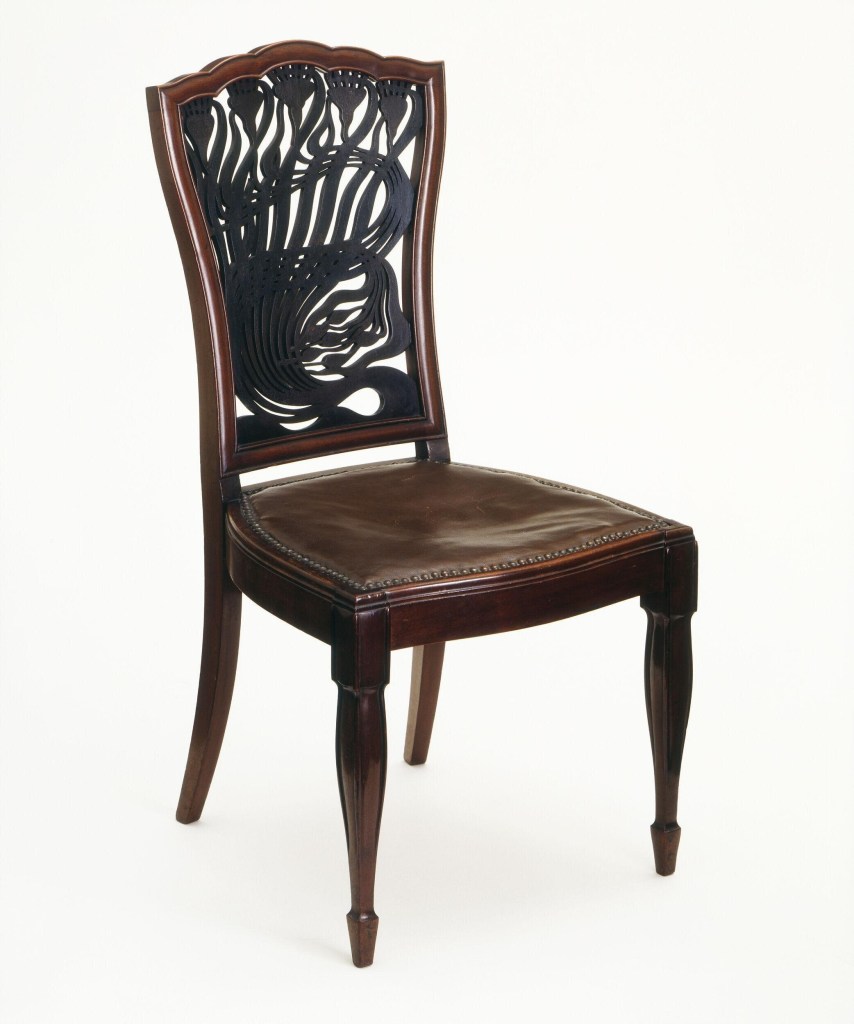 A mahogany chair designed by Arthur Heygate Mackmurdo in 1881, featuring an intricately carved and painted backrest with organic, flowing Art Nouveau motifs. The chair was likely made by Collinson and Lock and has a replacement green leather seat.