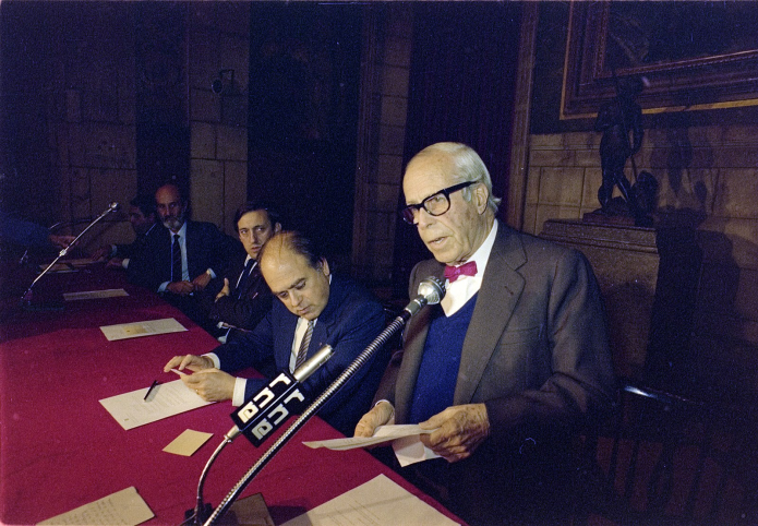 Josep Lluís Sert speaking at a conference, wearing a bow tie and glasses, with other officials seated at a table.