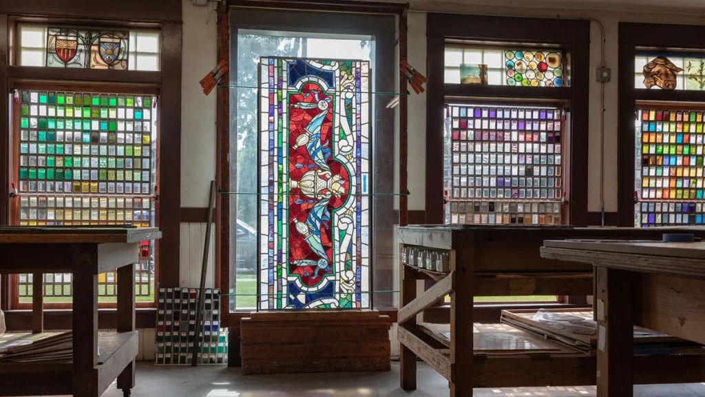 Interior of Judson Studios' stained glass workshop, featuring stained glass panels and colorful sample pieces displayed near large windows.

