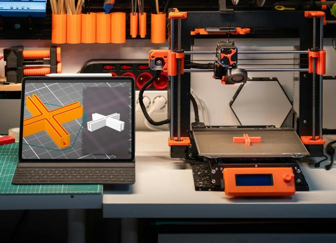 A laptop displaying a CAD design next to a 3D printer creating a prototype in a well-equipped industrial design workshop.