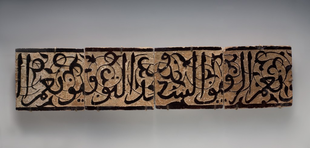 Panel of four Islamic calligraphic tiles with carved Arabic inscriptions on a dark glazed ground, 14th–early 15th century.